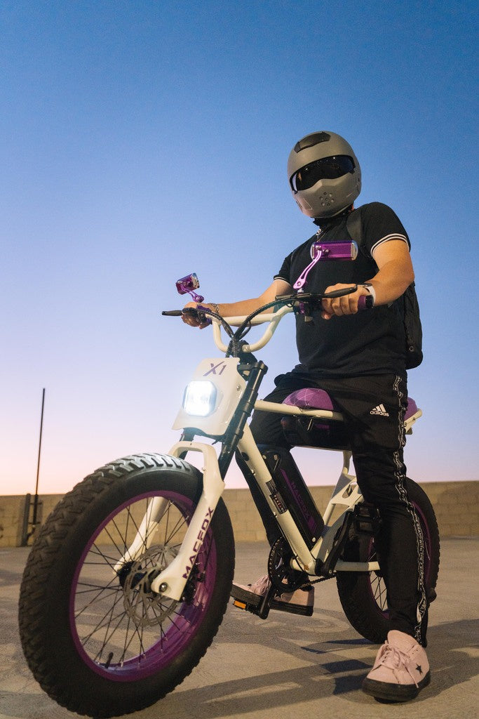 Rider wearing a helmet standing over a Macfox X1 electric bike at dusk, its bright rectangular headlight illuminating the area.