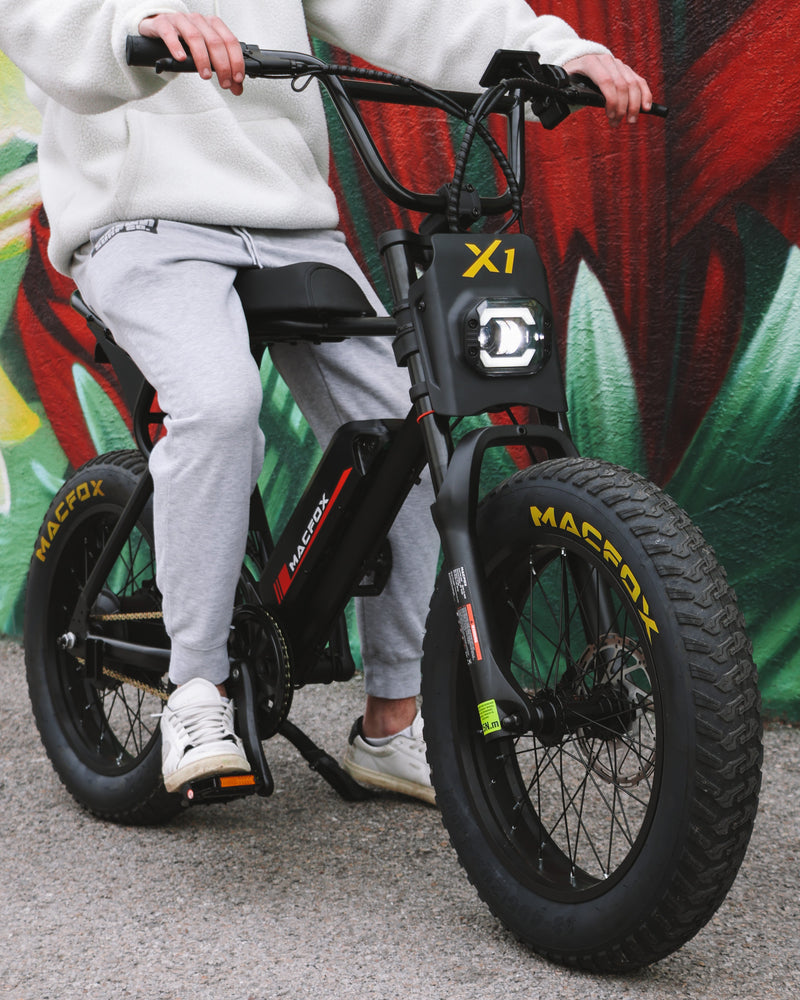 Close-up of a Macfox X1 electric bike with a rider preparing to pedal, showing chain, crankset, and drivetrain details.