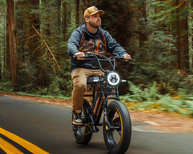 Person riding an electric bike through a forested area, with large tires and a front light, wearing a cap and casual clothing, surrounded by greenery on the road.