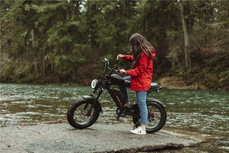 A person in a red jacket is standing next to an electric bike by a river, adjusting something on the bike while surrounded by trees.