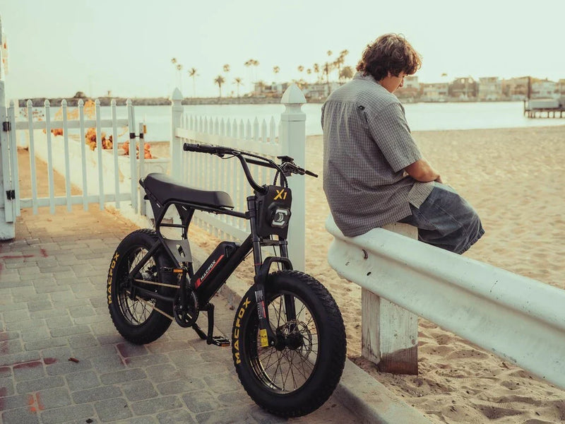 A young man is sitting on the railing on a Macfox X1S Commuter Electric Bicycle parked on the beach. 