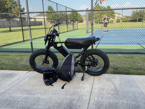A black Macfox electric bike parked beside a sports court with a helmet and backpack.