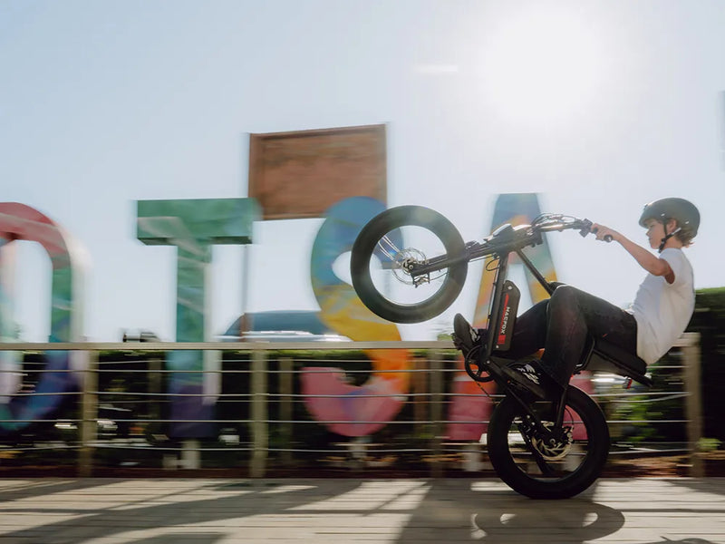 Rider performing a wheelie on a Macfox X1S commuter electric bike in front of colorful signs