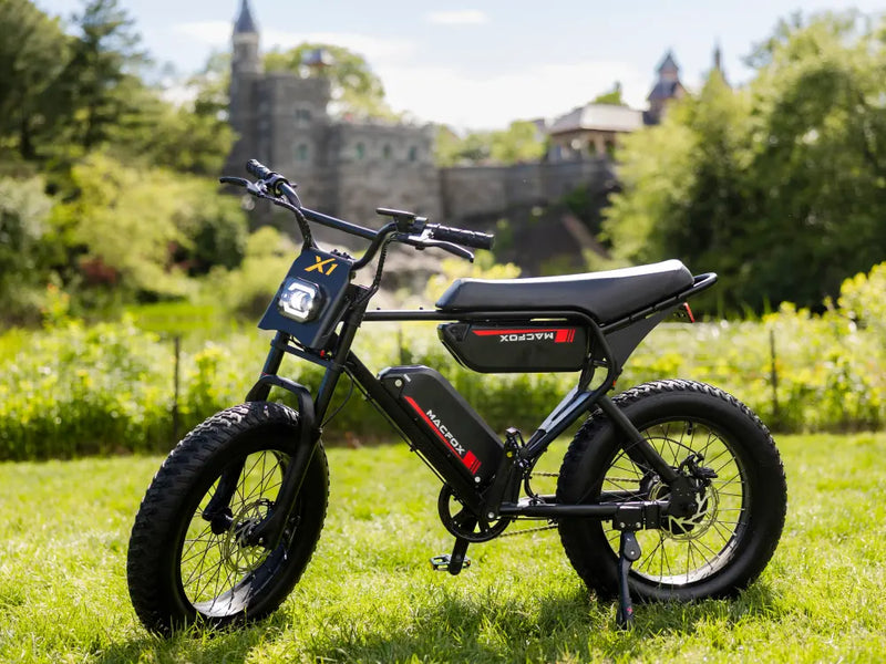 Macfox X1S electric bike parked on a sunny grassy trail with trees and a distant building in the background