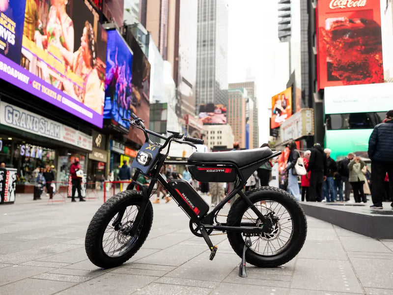 Macfox X1S electric bike parked in a busy city square surrounded by tall buildings and billboards